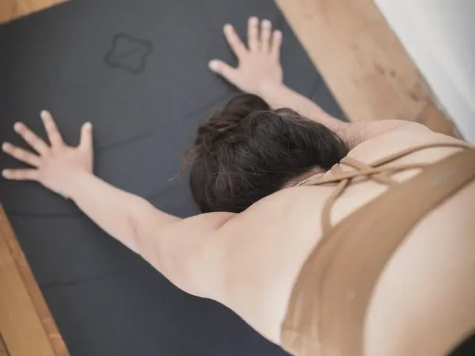 Une femme en posture de l'enfant sur un tapis de yoga, dans une pièce au parquet clair.