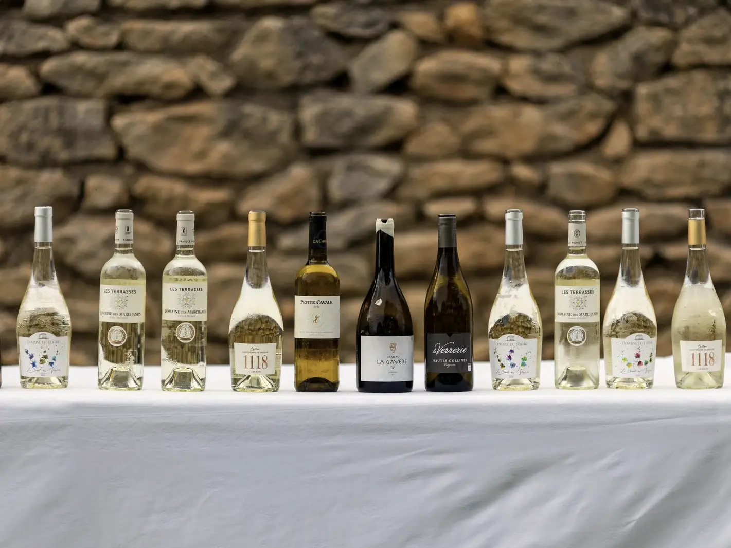Une rangée de bouteilles de vin alignées sur une nappe blanche, devant un mur de pierre.