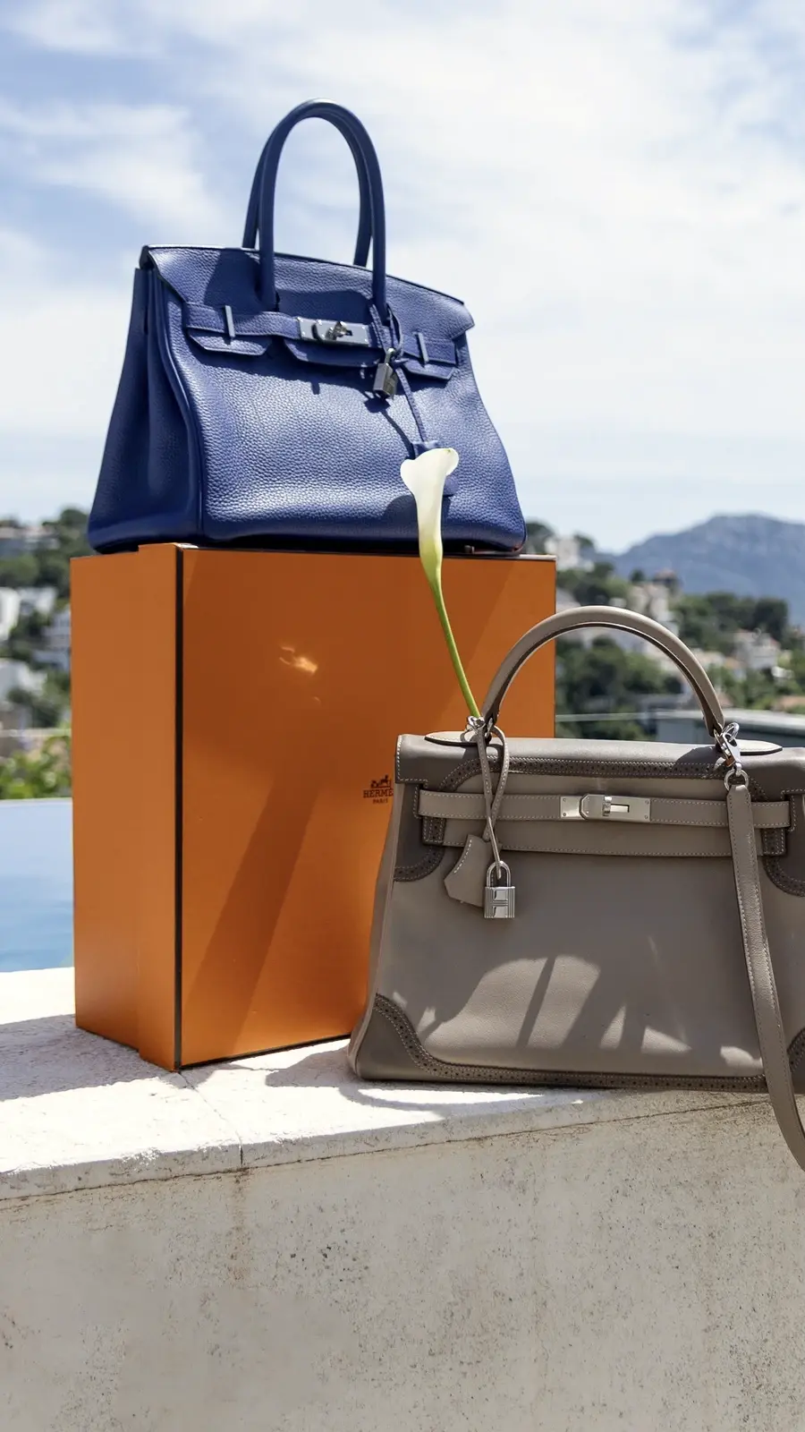 Deux sacs à main de luxe, un bleu et un gris, posés de part et d'autre de leur boîte orange, au bord d'une piscine face à la baie.