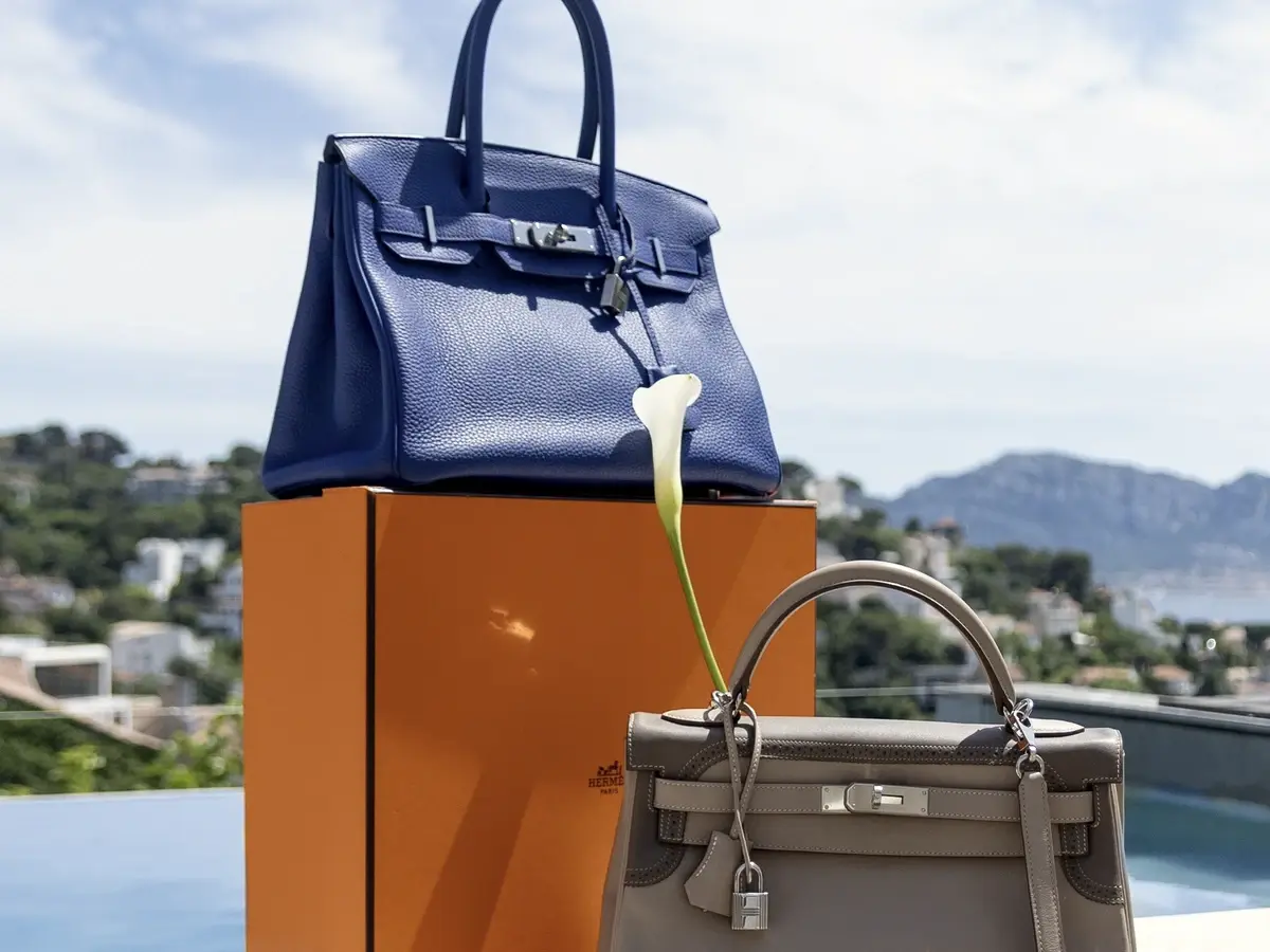 Deux sacs à main de luxe, un bleu et un gris, posés de part et d'autre de leur boîte orange, au bord d'une piscine face à la baie.